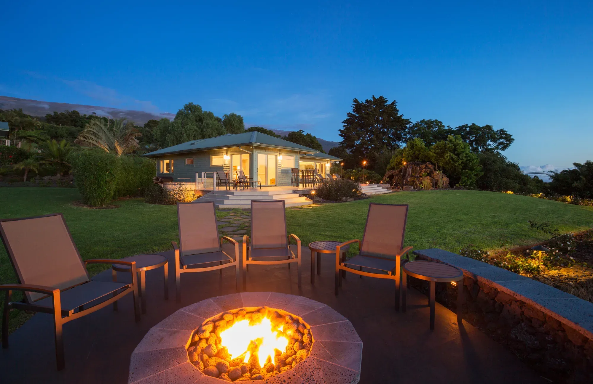 Outdoor lounge chairs around a built-in stone fire pit at dusk
