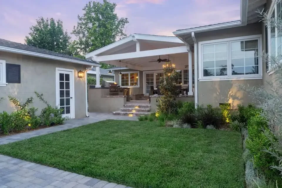 Covered outdoor living area with a pergola, outdoor kitchen and lawn at dusk